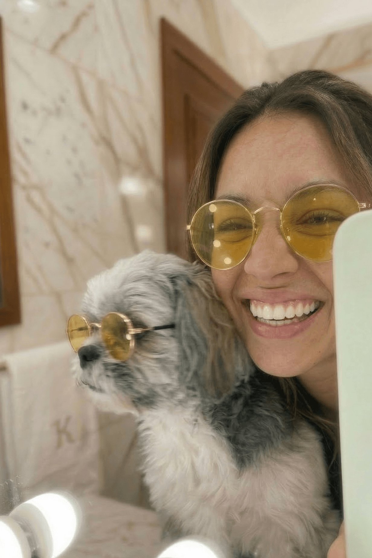 Woman holding a small dog wearing gold sunglasses in a bathroom setting